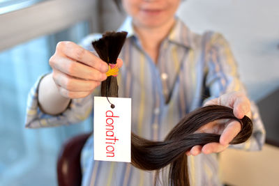 Long brown cut tail with tag in female hands, woman donation of haircut, depth of field