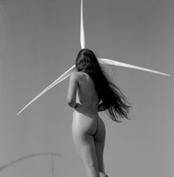 Rear view of naked woman standing by wind turbine against sky