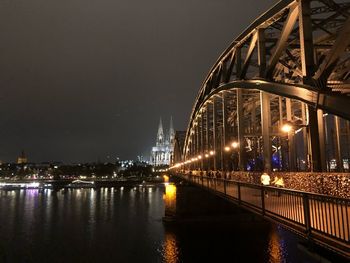 Illuminated bridge over river by buildings against sky at night