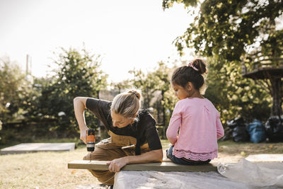 Daughter looking at mother using drill machine on wooden plank in backyard