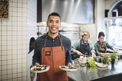 Portrait of smiling waiter holding fresh food plates against female chefs in restaurant kitchen