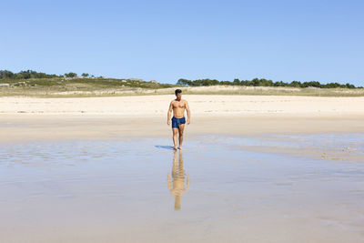 Full length of shirtless man standing on beach against clear sky