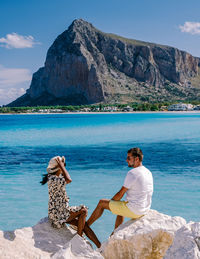 Rear view of woman sitting on rock by sea against mountain