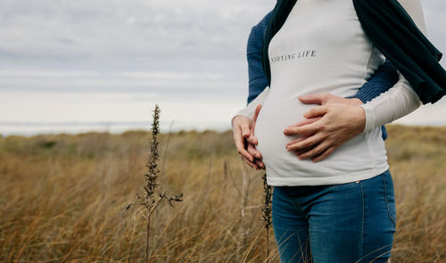 Midsection of pregnant couple touching abdomen while standing on grass against sky