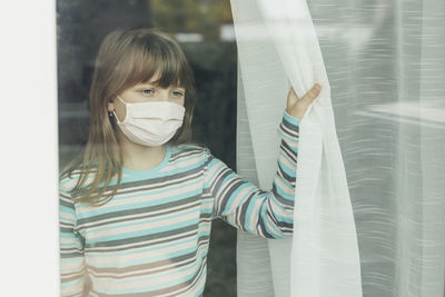 A school-age girl in a protective medical mask looks out the window. 