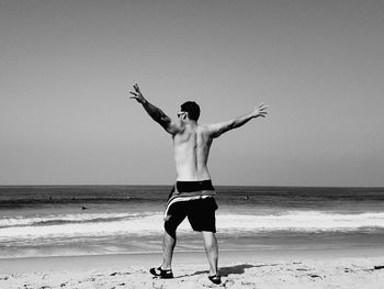 Rear view full length of shirtless man with arms outstretched at beach against sky