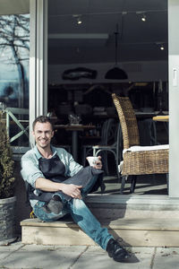 Portrait of confident owner with coffee cup sitting at entrance of cafe