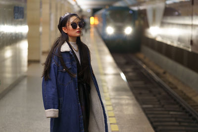 Stylish asian girl in glasses and street fashion outfit at underground platform with arriving train