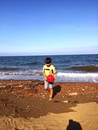 Boy carrying ball against sea at beach on sunny day