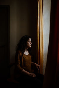 Thoughtful woman looking through window while standing in dark at home