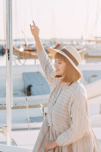 Happy young woman in dress and hat walks on the beach near the boats