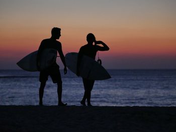 Silhouette friends with surfboards walking at beach against orange sky