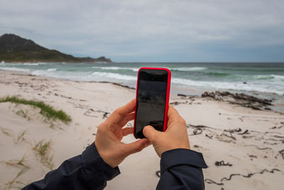 Midsection of person holding mobile phone at beach against sky