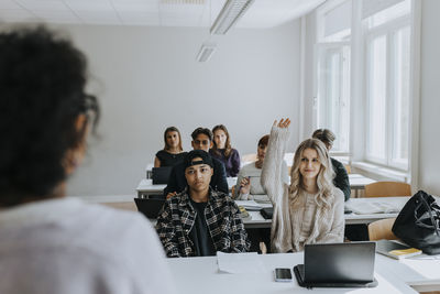 Young female student with hand raised sitting by friends looking at teacher in classroom