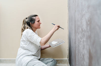 Blonde barefoot woman sitting on her floor starts painting a brown wall into a grey color with a small paintbrush. the woman wears a white shirt and a grey trousers. horizontal photo.