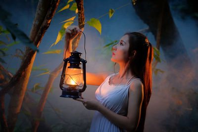 Close-up of young woman with illuminated lantern standing by tree