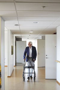 Full length of senior man looking away while walking with rollator in hospital corridor