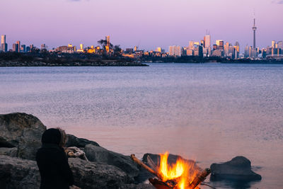 Woman sitting by campfire against river in city at dusk