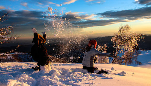 Kids throwing snow against cloudy sky during sunset