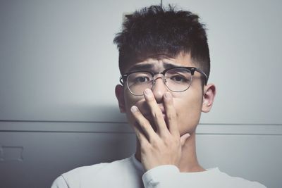 Portrait of man wearing eyeglasses covering mouth against wall