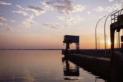 Scene of pier and crane and their reflection in the see under cloudy sky during sunset