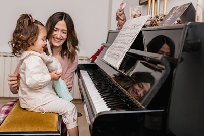 Cheerful mom interacting with charming girl holding toy against piano with music sheet in house