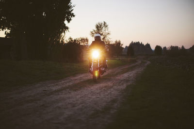 Male biker riding motorcycle on dirt road against clear sky during sunset