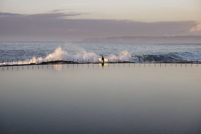 Distance view of man with surfboard at beach against sky during sunrise