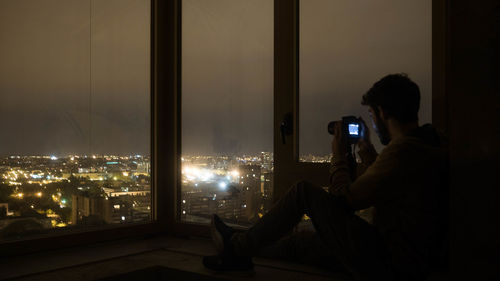 Man photographing illuminated cityscape while sitting by window at night