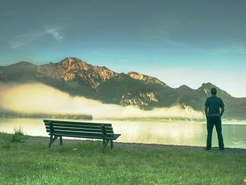 Rear view of man standing by lake against sky during sunset