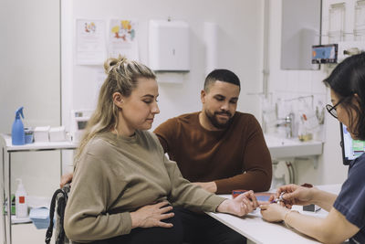 Gynecologist doing sugar test of pregnant woman sitting by man at desk in medical clinic