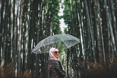 Smiling woman holding umbrella while standing against trees