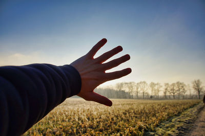 Cropped hand of man blocking sunbeam on field against sky