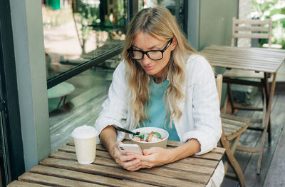 A modern woman sits on a terrace in a cafe having lunch and using her phone to surf the internet.