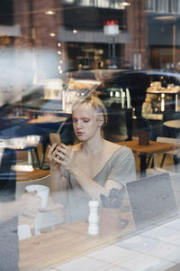 Young androgynous customer using smart phone while sitting in cafe seen from window