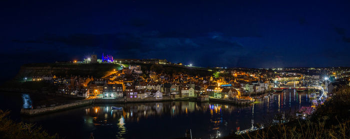 Illuminated buildings by river against sky at night