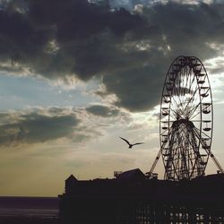 Low angle view of silhouette ferris wheel and bird against cloudy sky during sunset