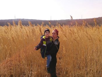 Woman carrying son while standing amidst crops on field against sky