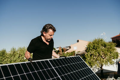 Mature technician man holding solar panels on house roof for self consumption energy. renewable