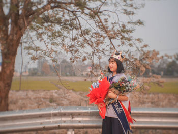 Side view of young woman holding bouquets standing against sky