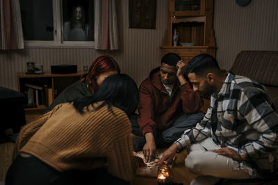 Multiracial friends touching ouija board while ghost seen through window in house