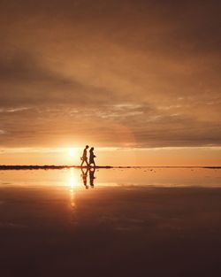 Silhouette man and woman walking on shore against cloudy sky during sunset