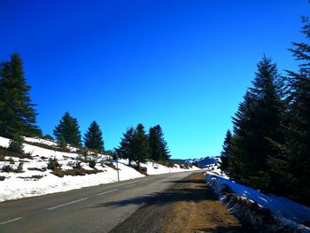 Road amidst trees and snowcapped mountains against clear blue sky