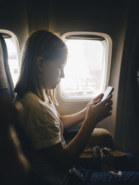 Side view of girl using mobile phone while sitting by airplane window