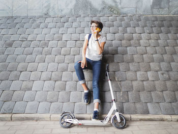 Smiling woman talks by smartphone after riding kick scooter. female on grey tiled wall background.