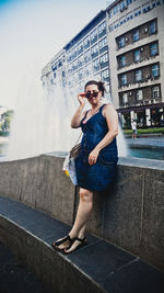 Woman wearing sunglasses standing by retaining wall against fountain in city