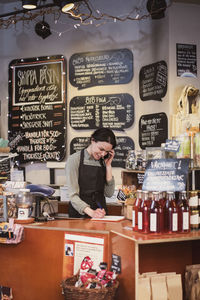 Smiling owner talking on phone while writing in note pad at checkout counter of store