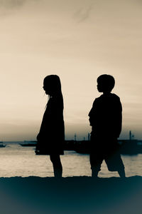 Silhouette boy and girl standing at beach against sky during sunset