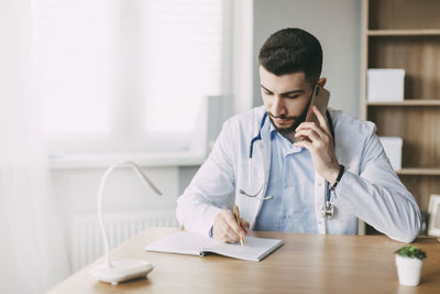 A young doctor of oriental appearance is talking on the phone, sitting at the table and making notes 