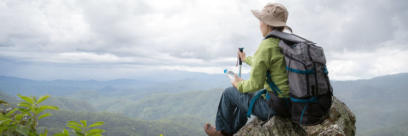 Banner panorama of young female tourist with backpack relaxing on top rock 